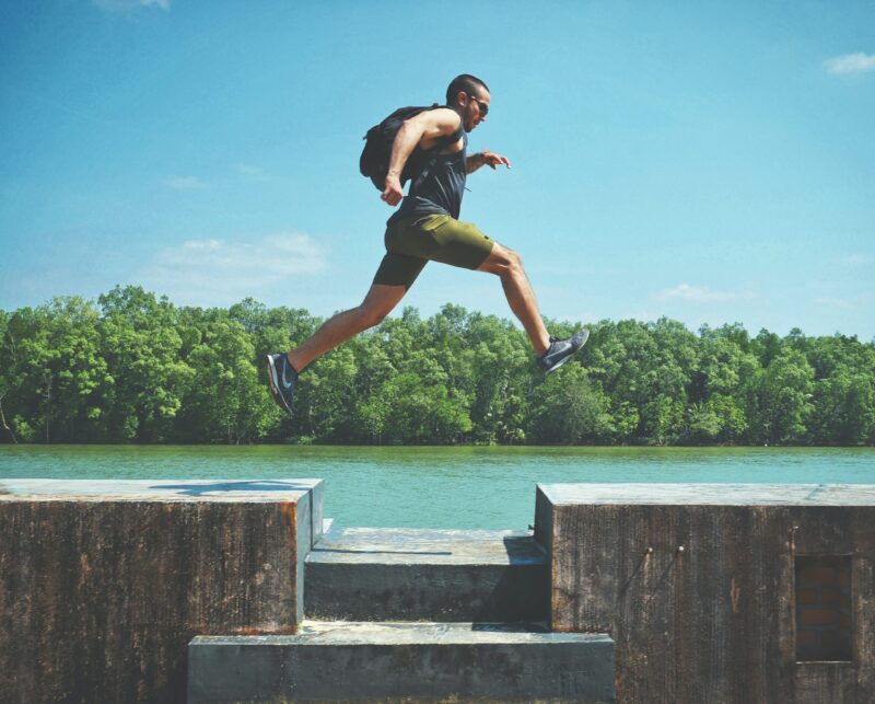 A man in athletic clothing wearing a backpack, who is jumping over a gap with a river and trees in the background.
