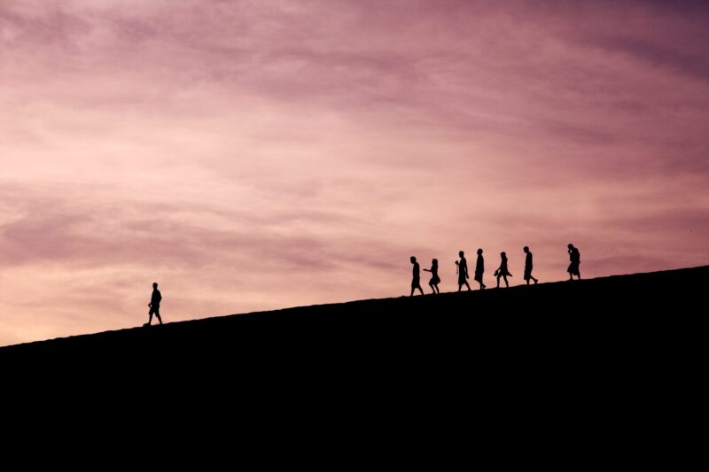 a group of people silhouetted against a pink sky, one man leading in front of the others