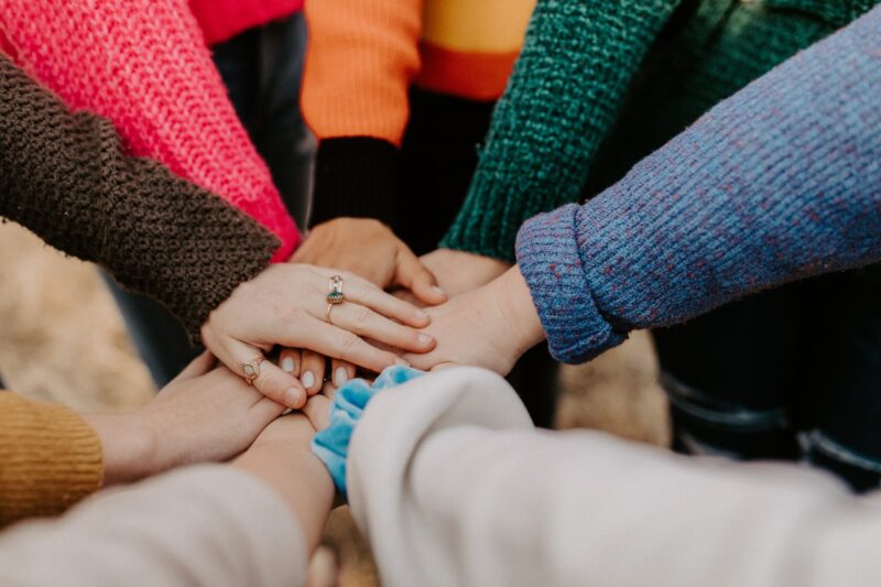 A photo of several peoples' hands stacked on top of each other in a circle, with colorful sweater sleeves, getting ready for a cheer