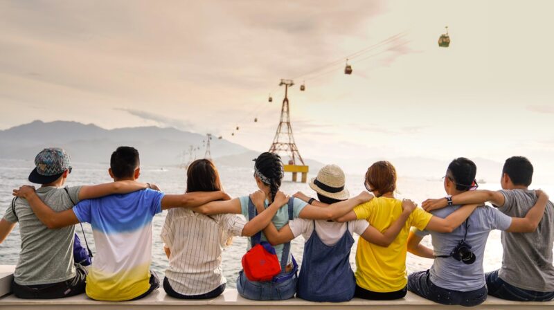 an image of eight people from behind sitting on a barrier near a body of water with their arms around each other