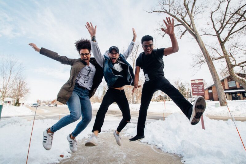 Three friends jumping from a snowy sidewalk into the air, excited with hands raised.